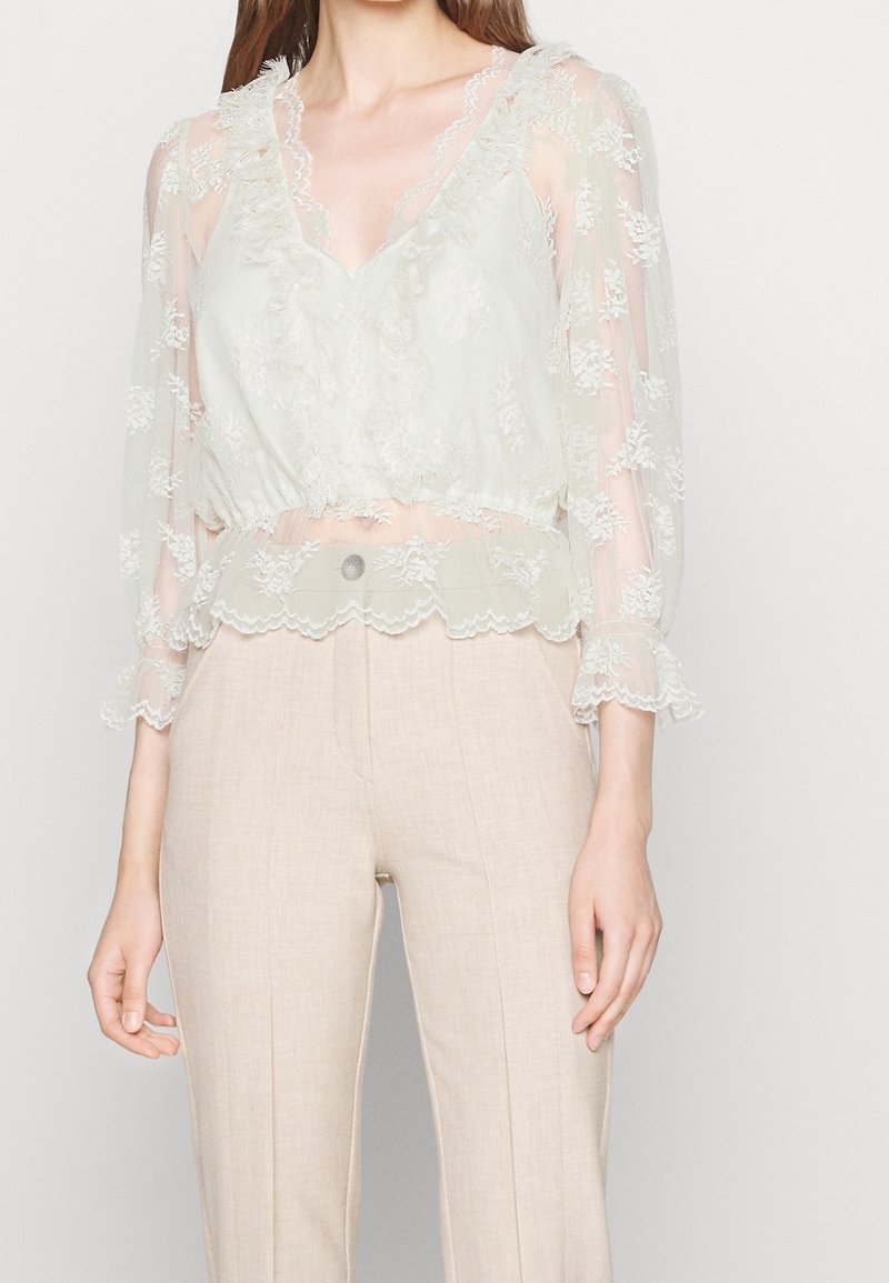 Blouse vert clair transparente avec des détails en dentelle, décolleté en V et manches volantées ; associée à un pantalon en lin beige taille haute.