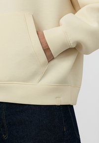 Sweat-shirt beige avec une poche kangourou, des poignets côtelés et une petite étiquette sur l'ourlet. Le tissu semble doux et texturé.