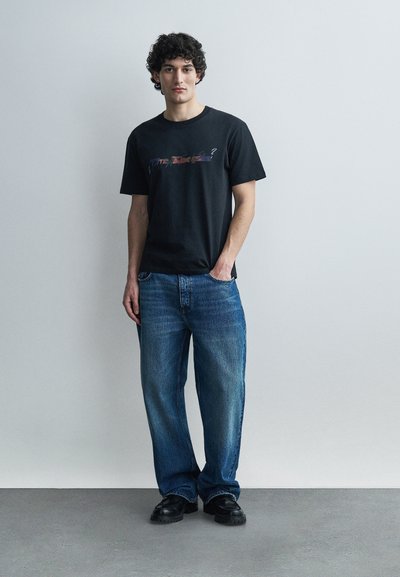 Νεαρός με κατσαρά μαλλιά στέκεται μπροστά σε γκρίζο τοίχο φορώντας μαύρο μπλουζάκι με σχέδιο, μπλε τζιν και μαύρα παπούτσια, με το ένα χέρι στην τσέπη.