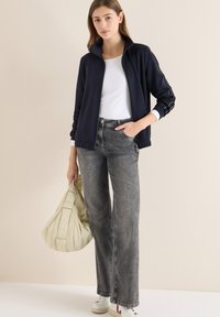 Granatowa kurtka zapinana na zamek błyskawiczny z białymi akcentami, biała koszulka, szare jeansy z szerokimi nogawkami i białe sneakersy, trzymająca beżową torebkę.