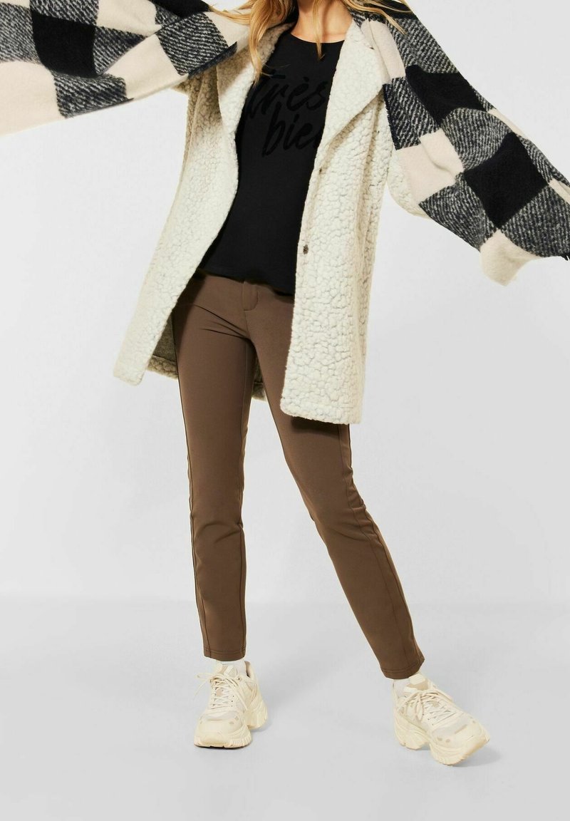 Femme portant un manteau en peau retournée beige sur une chemise noire, un pantalon slim marron, des baskets blanches épaisses, tenant une écharpe à carreaux noir et blanc.