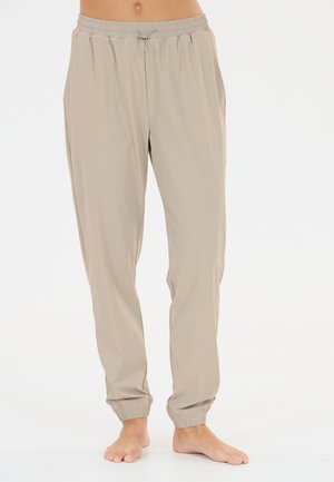 Pantaloni da jogging beige con vita elastica, coulisse e polsini alle caviglie, indossati a piedi nudi da una persona in piedi su sfondo bianco.