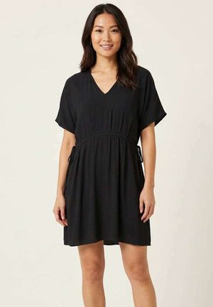 Femme debout et souriante, portant une robe noire courte à col en V, manches courtes et détails noués sur les côtés.