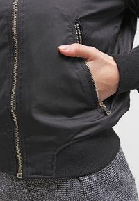 Main dans la poche zippée d'une veste noire avec une ceinture côtelée, portée sur un pantalon à motifs noir et blanc.