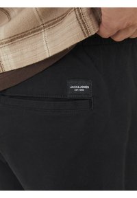 Svarta byxor med en bakficka som har en etikett med texten "JACK & JONES EST. 1990." Tyget verkar mjukt med en slät yta.