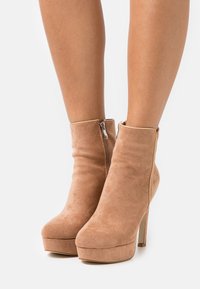 Bottines beige en daim avec une plateforme haute et un talon stiletto, dotées d'une fermeture éclair latérale et d'une texture lisse.