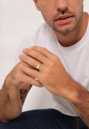 Gold metal ring with a sleek design, featuring a cross engraving. The model's hands are shown resting together on a plain background.