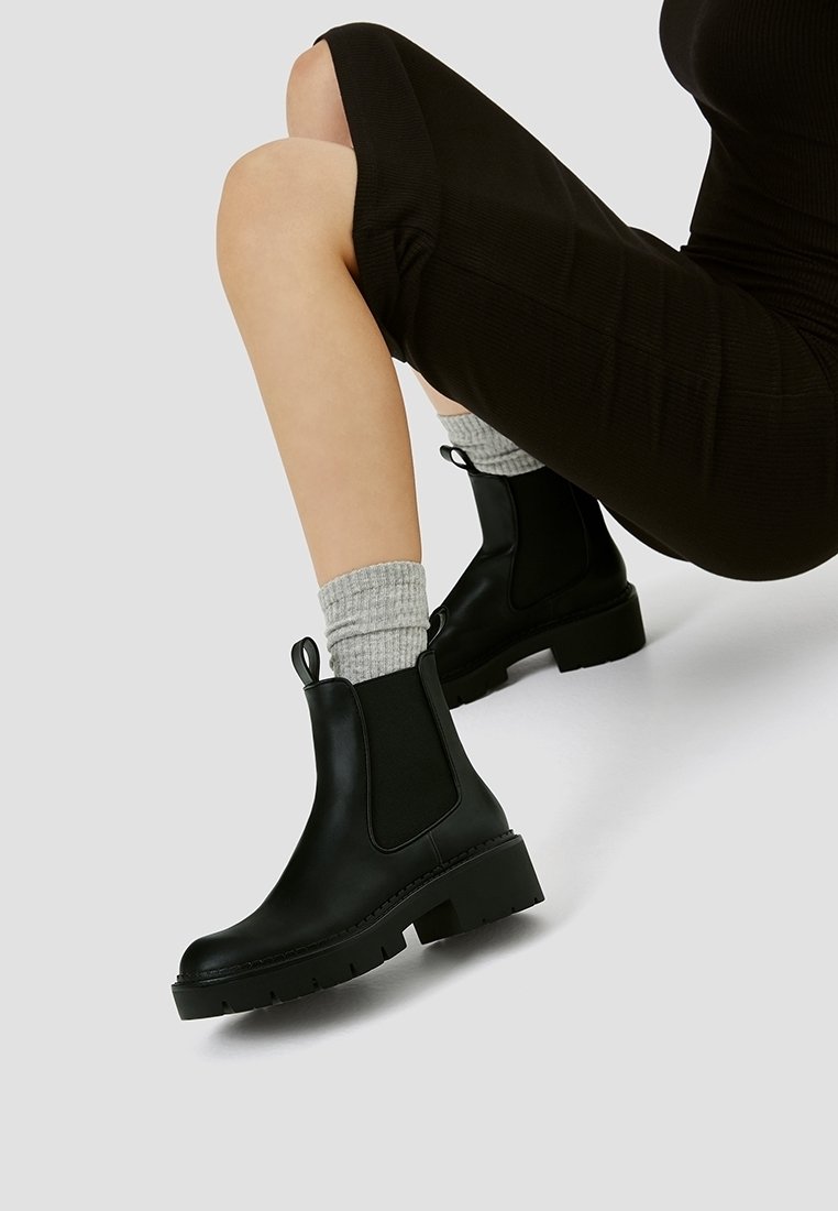 Bottes Chelsea noires avec panneaux latéraux élastiques, semelles en caoutchouc épaisses et languettes de tirage, portées avec des chaussettes grises et une robe noire.