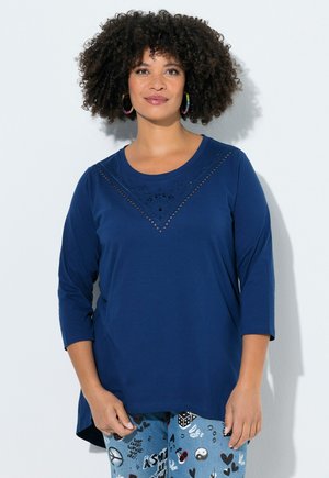 Kvinde med krøllet hår iført en blå bluse med lange ærmer med hulmønster og mønstrede jeans med symboler og tekst.