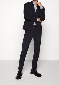 Donker marineblauw pak met een op maat gemaakte blazer, slim-fit broek en een blauw-wit gestreefd overhemd, gecombineerd met glanzende zwarte dresschoenen.