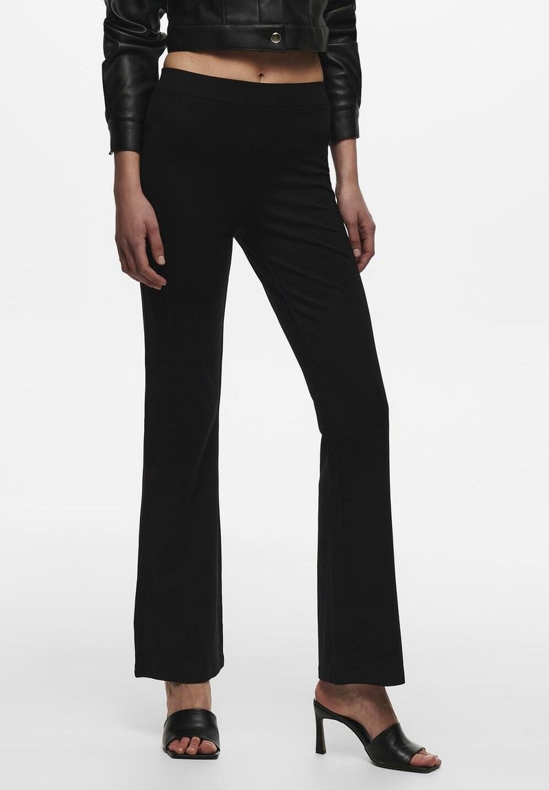 Femme portant un pantalon évasé noir taille haute, une veste courte en cuir noir et des mules noires à talons hauts et bouts ouverts, debout sur fond blanc.
