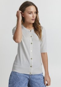 Kortærmet hvid cardigan lavet af blødt stof, med knaplukning foran og ribbet kant. Uden mønstre; minimalistisk design.