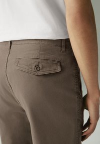 Brown cotton pants with a back pocket featuring a button closure, smooth texture, and a fitted design. Worn with a white shirt.