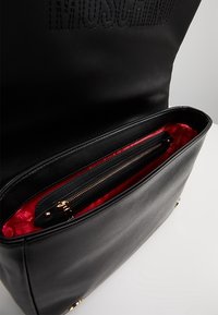 Czarna skórzana torebka z wytłoczonym logo, czerwoną podszewką, jedną kieszenią na zamek błyskawiczny oraz złotymi akcentami.