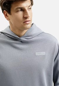 Jasnoszara bluza z kapturem z centralnie umieszczoną naszywką z logo, miękką teksturą oraz luźnym krojem z dużym, zwisającym kapturem.