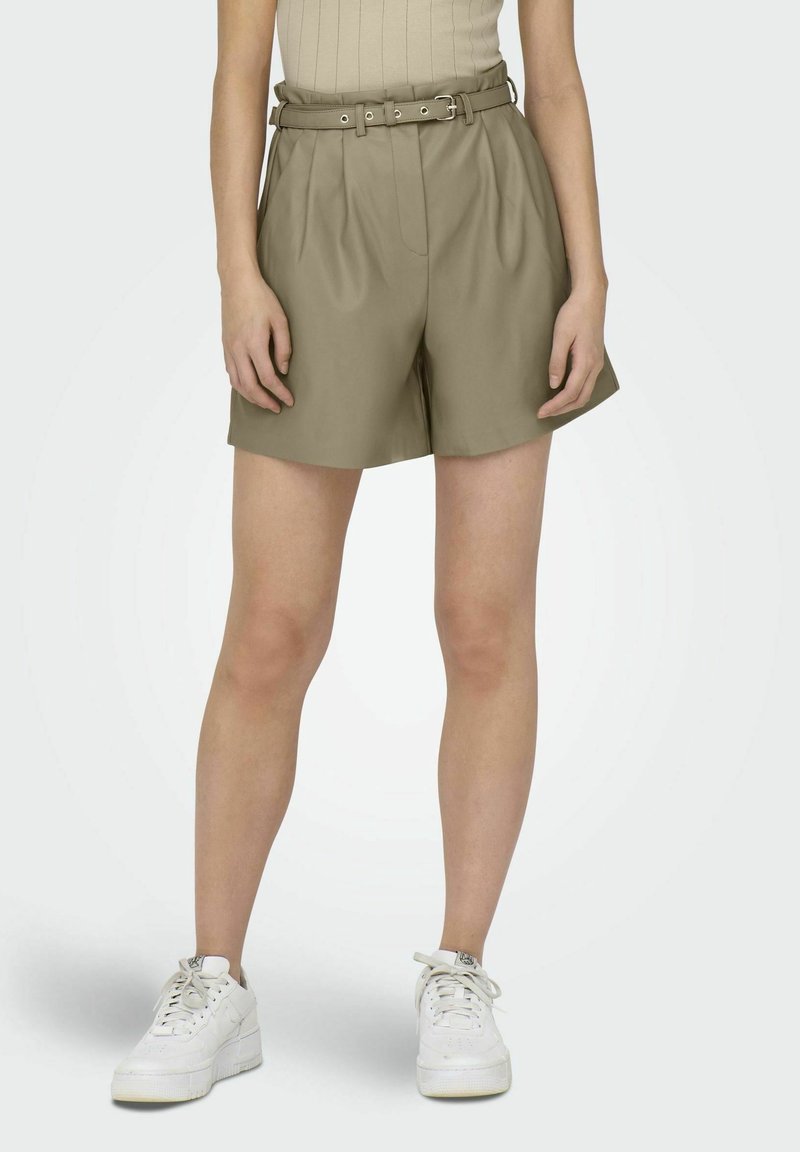 Jambes fines portant un short plissé beige taille haute avec ceinture, associé à des baskets blanches sur un fond clair uni.