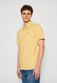 Žlté polo tričko s límcom, krátkymi rukávmi a malým vyšitým logom na ľavej hrudi. Tkanina sa zdá byť mäkká a hladká.