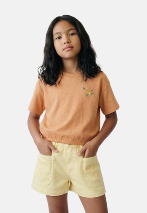 Jeune fille aux longs cheveux foncés portant un t-shirt orange avec une broderie florale et un short jaune avec des poches avant, mains dans les poches.