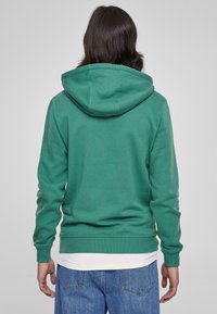 Felpa verde con cappuccio, dal tessuto morbido e con tasca a kangaroo. Presenta una vestibilità comoda e polsini a coste. Indossata sopra una t-shirt bianca.