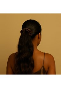 Brązowa satynowa gumka do włosów, która trzyma długie, falowane, ciemne włosy o gładkiej fakturze. Tło ma miękki beżowy odcień. Włosy związane w niski kucyk.