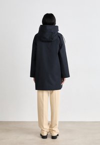 Tyrkysově modrá nepromokavá bunda s kapucí, rovný střih a elastické manžety. V kombinaci s béžovými kalhotami s širokými nohavicemi. Minimalistický design.