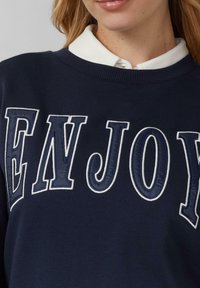 Sudadera de color navy con el texto "ENJOY" bordado en blanco. Presenta un cuello redondo acanalado y un color blanco en capas visible debajo.
