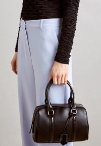 Sac à main en cuir noir avec finition brillante, poignées supérieures doubles, fermeture zippée et un petit pendentif. Porté avec un pantalon bleu clair et un haut noir texturé.