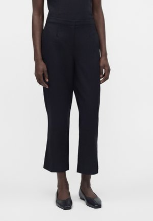 Personne portant un pantalon noir court à jambes droites et des chaussures noires à talon bas et bout carré, debout devant un fond uni.