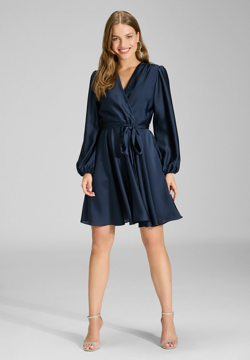 Marineblauwe wikkeljurk met lange pofmouwen en een ceintuur in de taille. Satijnen textuur met een uitlopende rok die boven de knie valt.