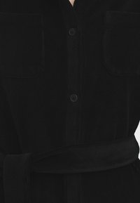 Abito camicia in velluto a coste nero con bottoni frontali, due tasche sul petto e una cintura coordinata annodata in vita.