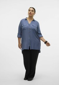 Blouse bleue à manches longues avec fermeture à boutons et manches retroussées, dotée de deux poches poitrine et d'un ourlet légèrement courbé, associée à un pantalon noir.