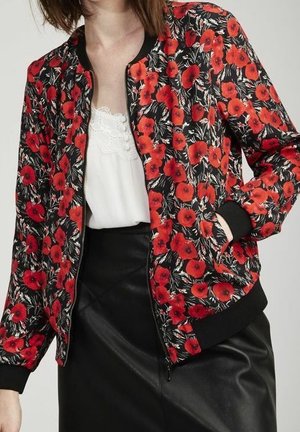 Kobieta w czarnej kurtce bomber z czerwonym nadrukiem maków na tle białej, koronkowej bluzki oraz czarnej skórzanej spódnicy, z ręką w kieszeni.