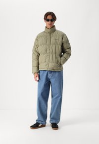 Puffjakke i olivengrønn med høy krage og quiltet tekstur. Brukt over vide blå jeans og svarte joggesko. Elegant design.