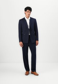 Sacou bleumarin, cu două rânduri de nasturi, cu accente din satin, purtat cu o cămașă albă și pantaloni bleumarin asortați, terminat cu încălțăminte din piele maro.