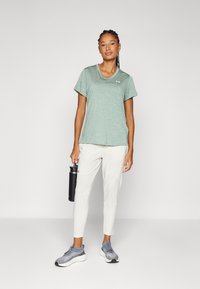 Camiseta deportiva de color verde claro con mangas cortas, escote en V, textura sutil, combinada con pantalones blancos ajustados y zapatillas de correr grises.