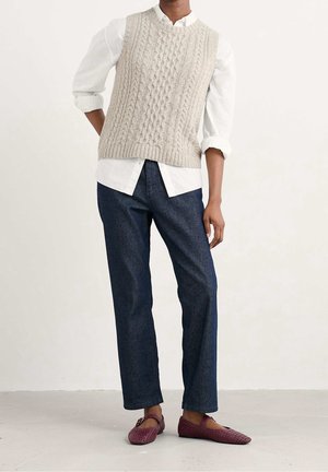 Personne portant une chemise blanche retroussée sous un gilet beige en maille torsadée, un jean bleu foncé et des chaussures plates tissées bordeaux.