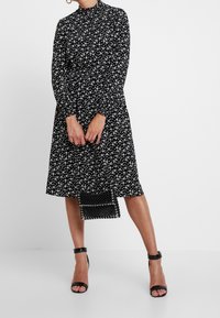 Svart klänning med vitt blommönster, hög hals, långa ärmar och avslappnad passform. Kombinerad med en liten svart pärlpåse och svarta klacksandaler.