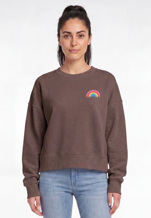 Frau mit dunklen Haaren im Pferdeschwanz trägt ein braunes Sweatshirt mit kleinem mehrfarbigem Regenbogen und dem Text "Women Power" vorne, kombiniert mit blauen Jeans.