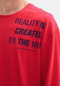 T-shirt en coton rouge à col rond, avec un imprimé graphique en texte bleu foncé qui dit "LA RÉALITÉ EST CRÉÉE PAR L'ESPRIT" et le logo de la marque.
