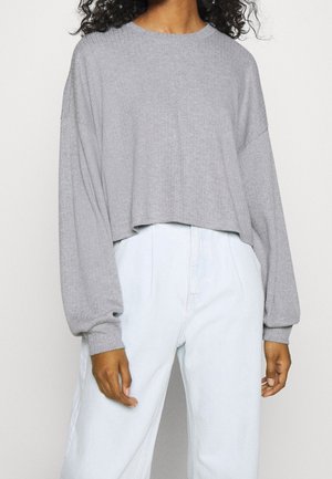 Osoba ubrana w jasnoszary, prążkowany, krótki sweter z długim rękawem oraz białe spodnie z wysokim stanem na jednolitym tle.