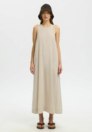 Frau trägt ein ärmelloses beiges Maxikleid, braunen gestrickten Fischerhut und dunkle gepolsterte Sandalen, steht vor einem schlichten weißen Hintergrund.