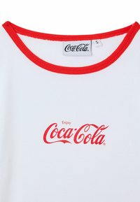 T-shirt bianco in cotone con scollatura rossa, caratterizzato da una grafica centrale rossa "Enjoy Coca-Cola" in un font stilizzato. Etichetta della taglia visibile.