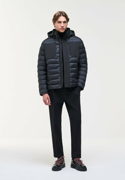 Un modèle masculin se tient devant un fond clair uni, vêtu d’une veste matelassée noire à capuche, d’un pantalon noir et de chaussures noires à lacets rouges.