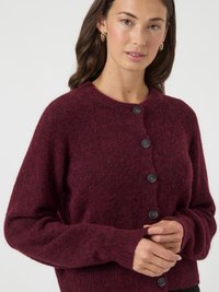 Mladá žena v bordovom plyšovom kardigáne s gombíkmi, s prekríženými rukami pred sebou, zlatými kruhovými náušnicami a prirodzeným líčením.