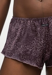 Κοντινό πλάνο ενός κορμού με σκούρο δέρμα που φοράει μωβ σορτς Calvin Klein με λεοπάρ σχέδιο και ελαστική μέση, σε λευκό φόντο.