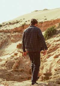 Homme portant des vêtements sombres et des lunettes de soleil marchant sur un terrain rocheux avec des collines de sable et une végétation clairsemée en arrière-plan.