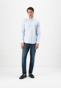 Mann som står mot en hvit vegg iført lyseblå skjorte, blå jeans og svarte loafers med dusk, ser på kamera.