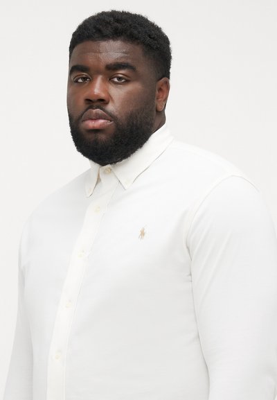 Chemise blanche à col, avec un petit logo doré sur la poitrine gauche et des manches longues avec poignets.