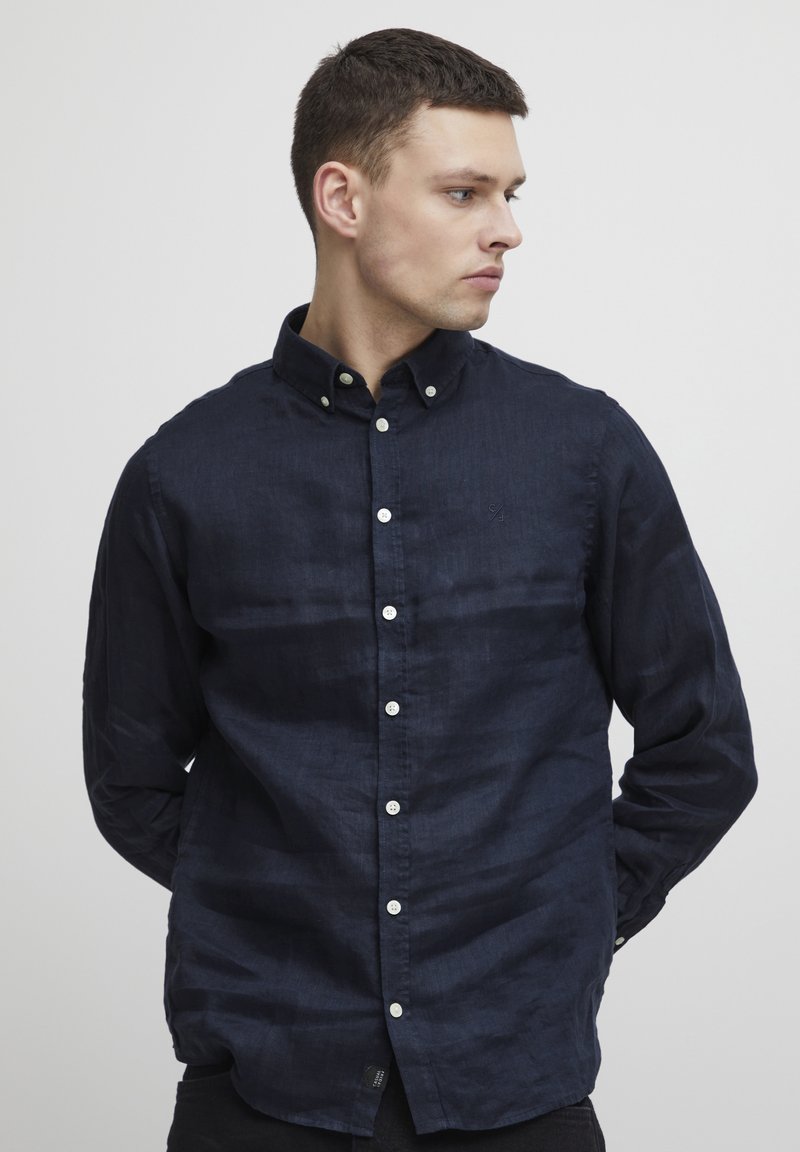 Camisa de botonadura azul marino en tejido ligero, con un cuello clásico, mangas de longitud completa y botones blancos en la parte delantera.