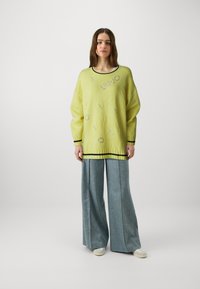 Suéter amarelo de malha com padrão de logotipo relevo, com acabamento em preto em contraste, combinado com calças de ganga de perna larga em azul claro e ténis brancos.